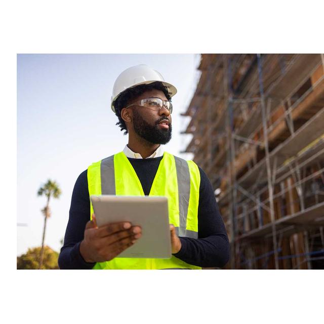 A man in a hardhat and safety vest holds a tablet and looks with concentration at a building under construction.